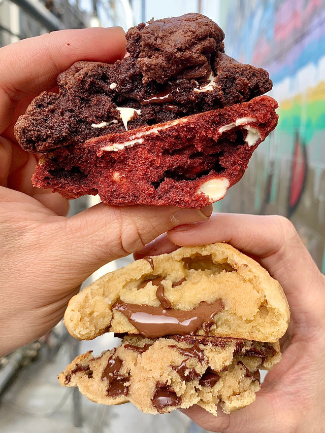 hands holding gourmet cookies including white chocolate chip, red velvet, and chocolate chip